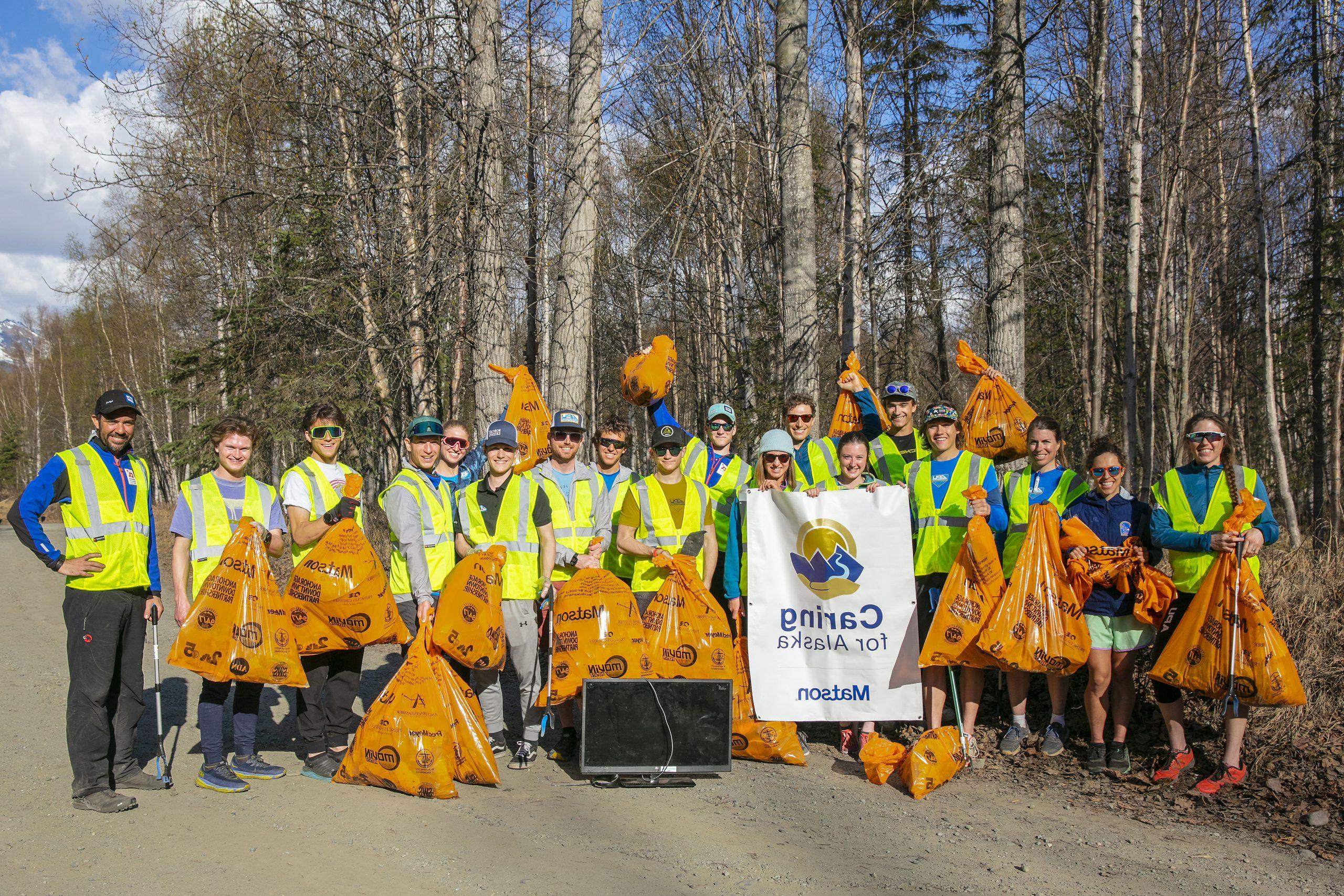 身穿黄色安全背心的志愿者拿着满满的橙色垃圾袋和“关爱阿拉斯加”横幅合影留念.