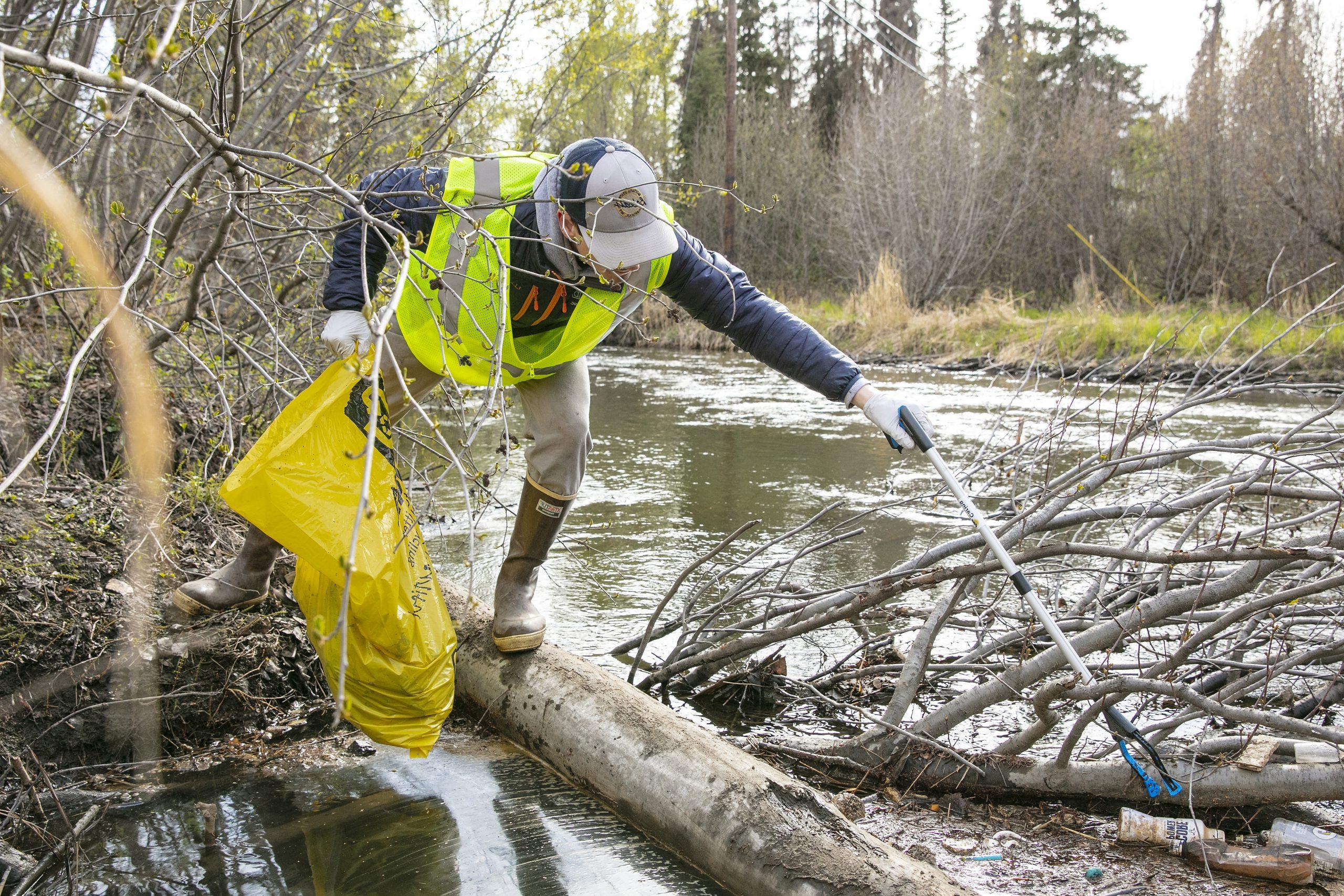 一名穿着橡胶靴和黄色安全背心的志愿者拿着一个黄色垃圾袋，拿着一个抓取器在坎贝尔溪捡垃圾.